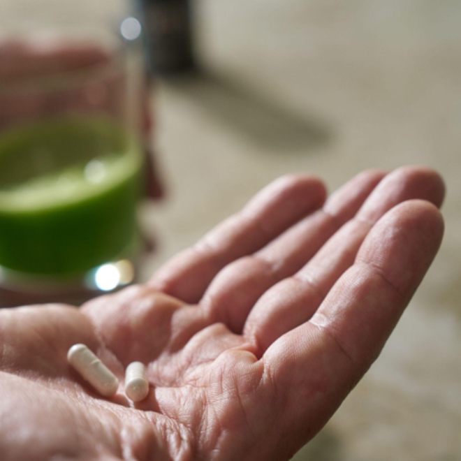 Two almä capsules resting in an open palm beside a glass of green aloe juice